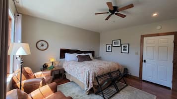 Une chambre à coucher comprenant un lit, une table de chevet, une lampe, une chaise, un ventilateur de plafond et une porte.