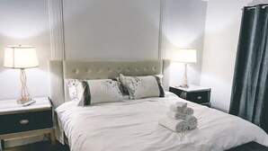 Een netjes opgemaakt bed met witte lakens en opgerolde handdoeken, twee nachtkastjes met lampen, en een donker gordijn voor het raam.