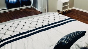 A bedroom with a bed, a white door, a small white nightstand, and a window with blue curtains.