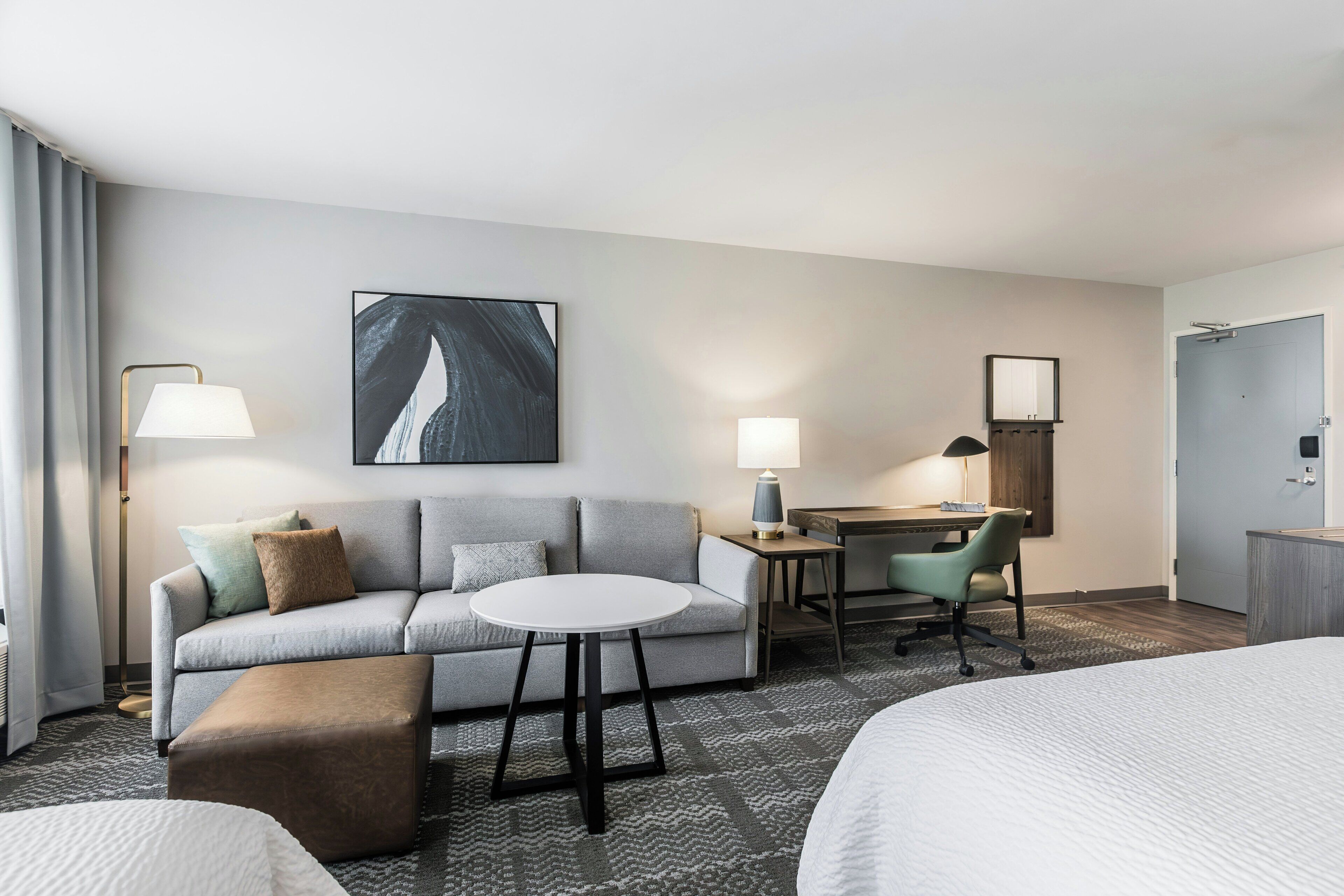 A hotel room with a sofa, a small round table, a desk, a chair, a bed, and a large painting on the wall.