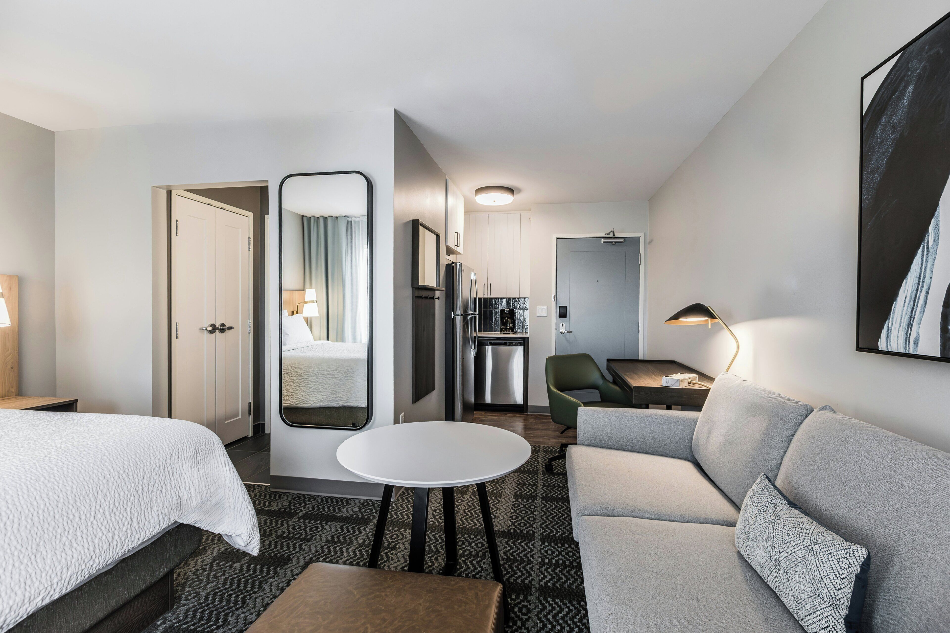 A hotel room with a bed, a seating area with a sofa and armchair, a small table, a mirror, and a kitchenette.