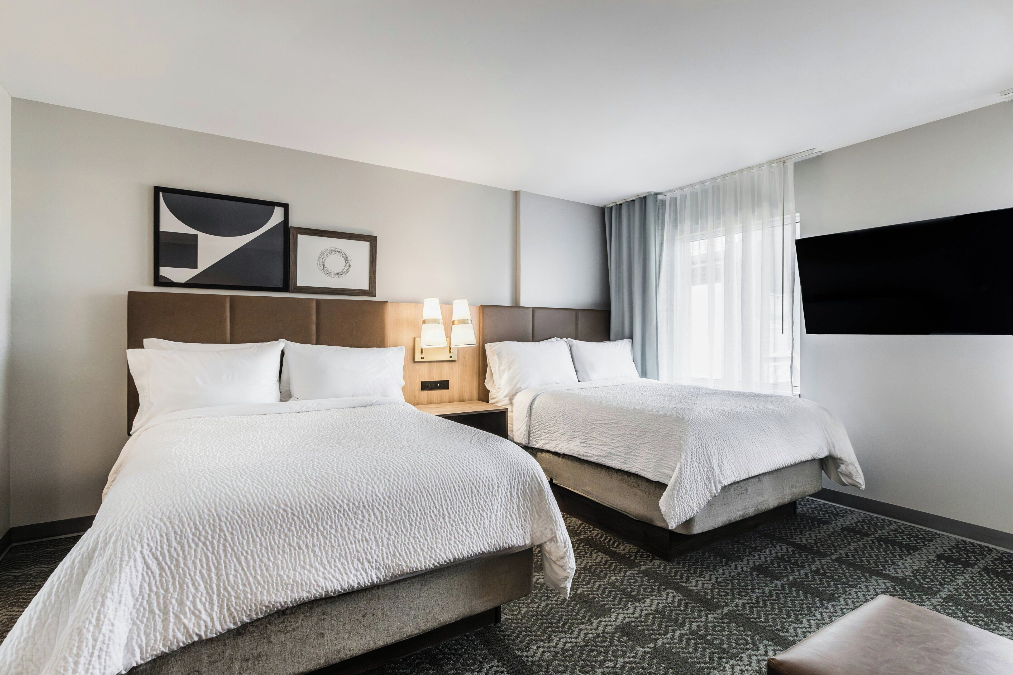 A hotel room with two beds, a nightstand with lamps, a large window with curtains, and artwork on the wall.