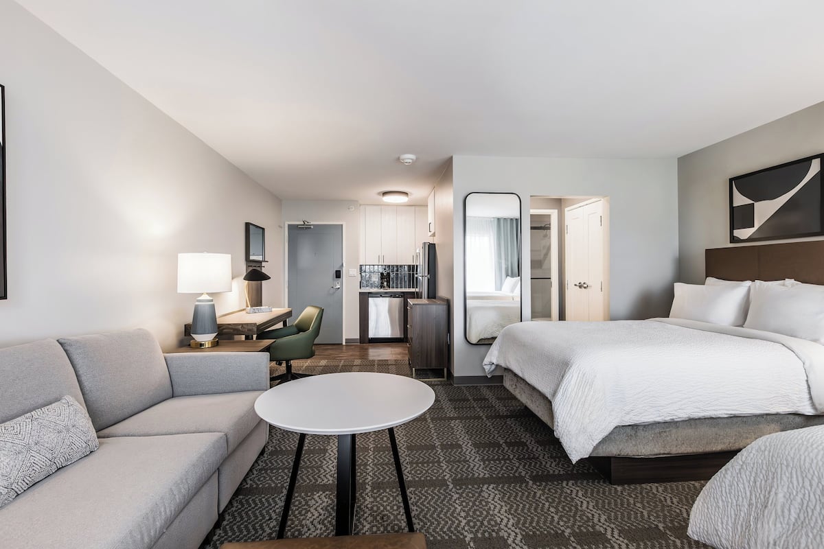 Ein Hotelzimmer mit Bett, einer Couch, einem kleinen Tisch, einem Schreibtisch, einem Stuhl, einem Spiegel und einer Küchenzeile.