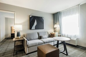 Et hotelværelse med en grå sofa, et rundt bord, en lampe, et billede og et vindue med gardiner.