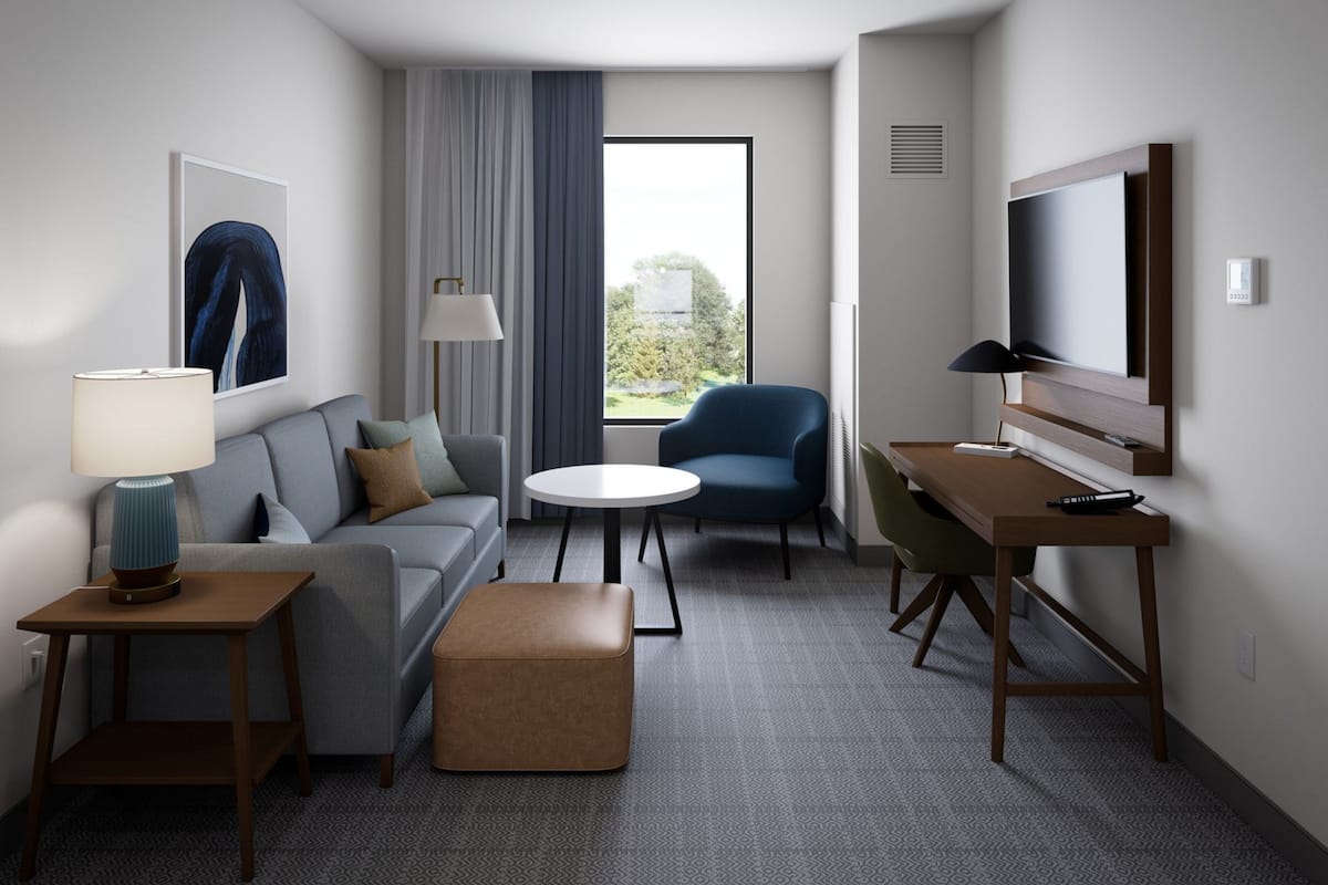 Ein modernes Hotelzimmer mit einer Couch, einem runden Tisch, einem blauen Sessel, einem Schreibtisch und einem an der Wand montierten Fernseher.