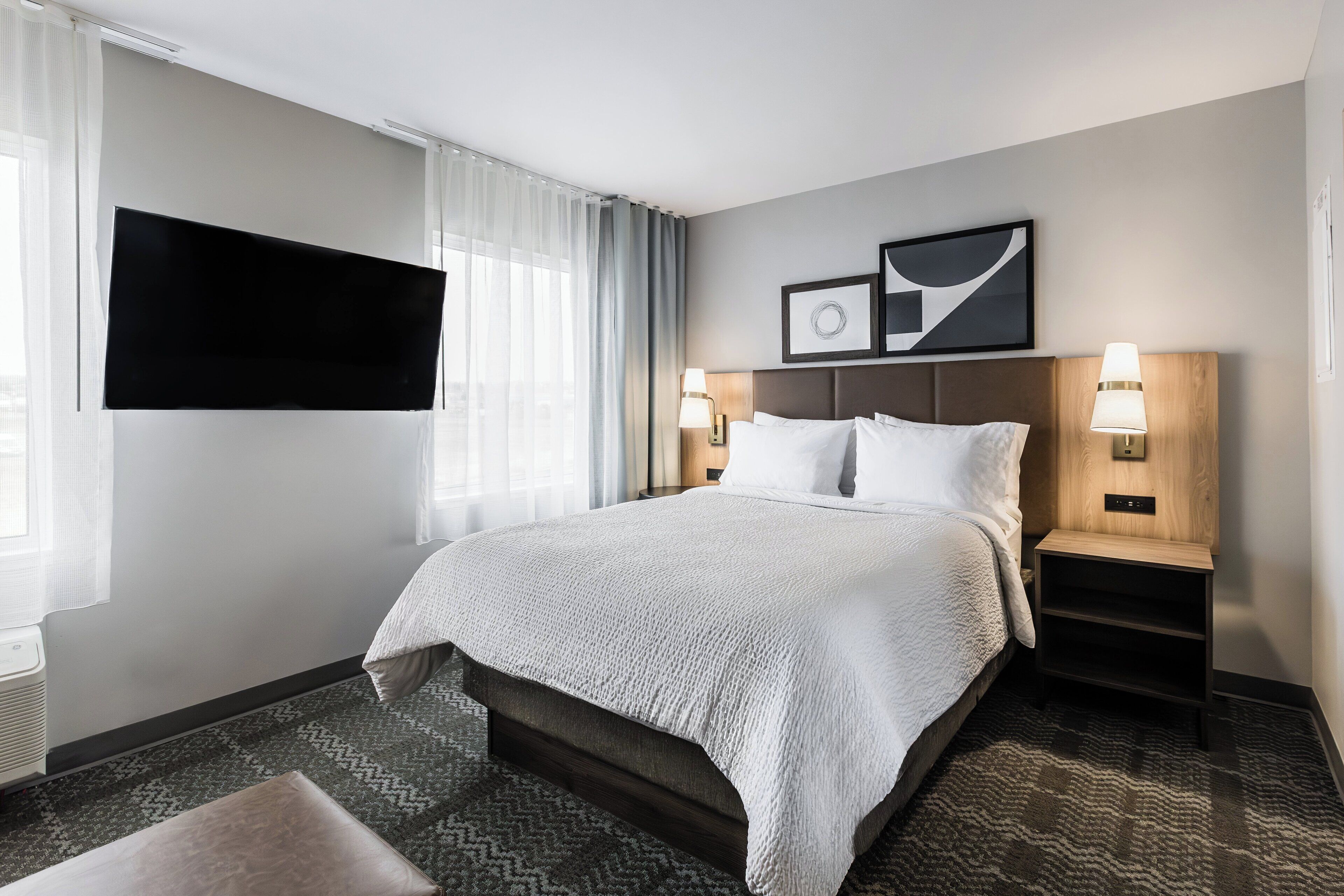 Une chambre d’hôtel avec un grand lit, un téléviseur à écran plat fixé au mur, une table de chevet avec une lampe et un tableau encadré au-dessus du lit.