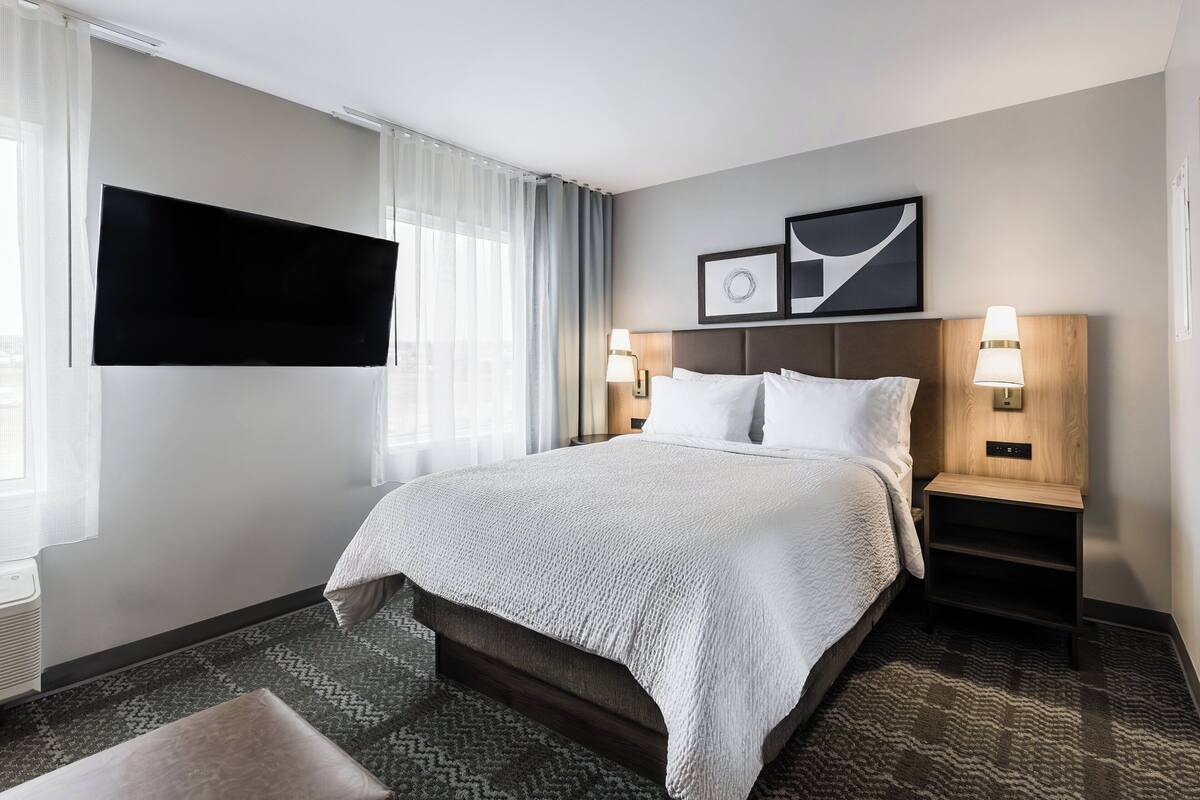 Ein Hotelzimmer mit einem großen Bett, einem flachbildfernseher an der Wand, einem Nachttisch mit Lampe und einem gerahmten Bild über dem Bett.