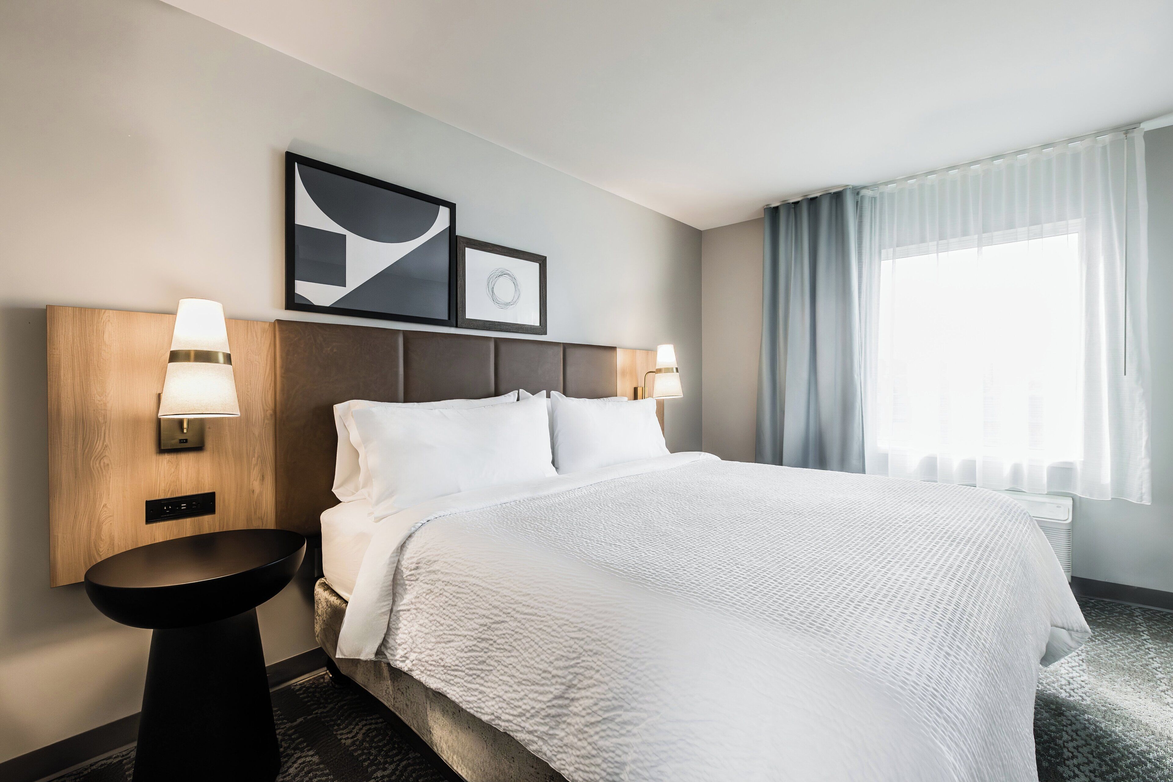 Une chambre d’hôtel avec un grand lit, deux lampes de chevet, une tête de lit, une œuvre d’art accrochée au mur et une fenêtre avec des rideaux.
