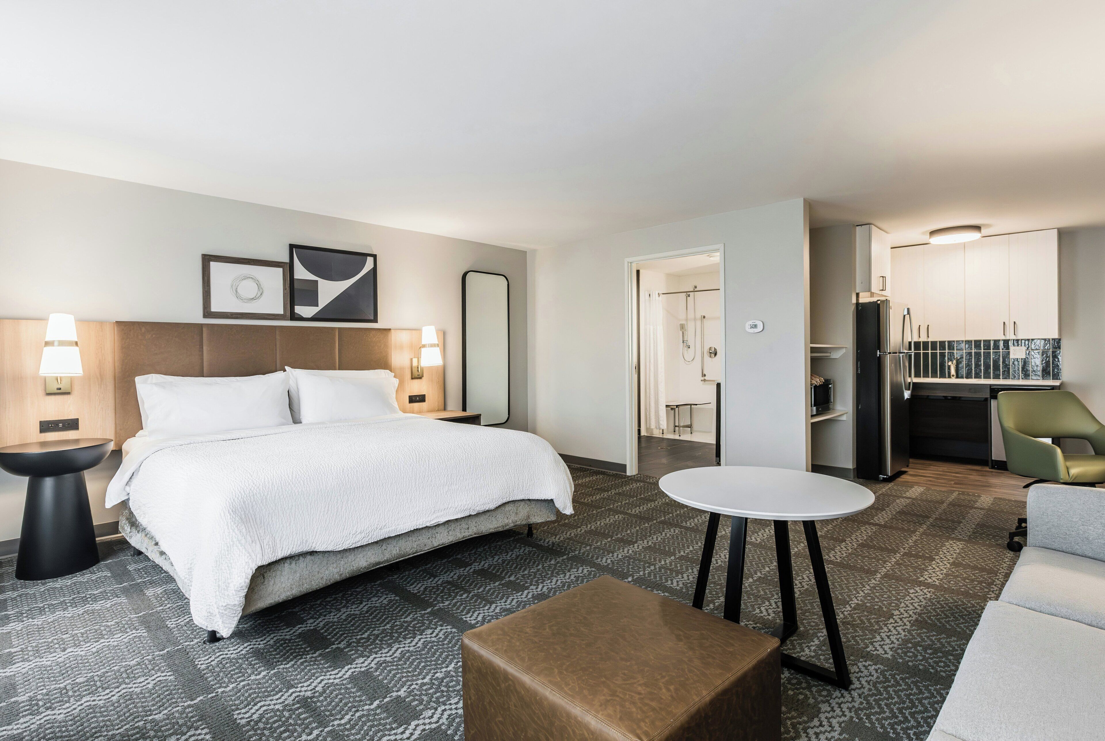 A hotel room with a bed, bedside tables, a round table, a brown ottoman, a shower area, and a kitchenette.