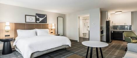 A hotel room with a bed, bedside tables, a round table, a brown ottoman, a shower area, and a kitchenette.