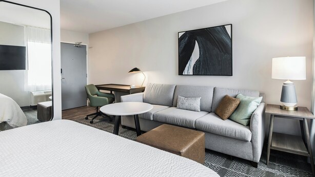 A hotel room with a bed, a sofa, a small round table, a lamp, and a painting on the wall.