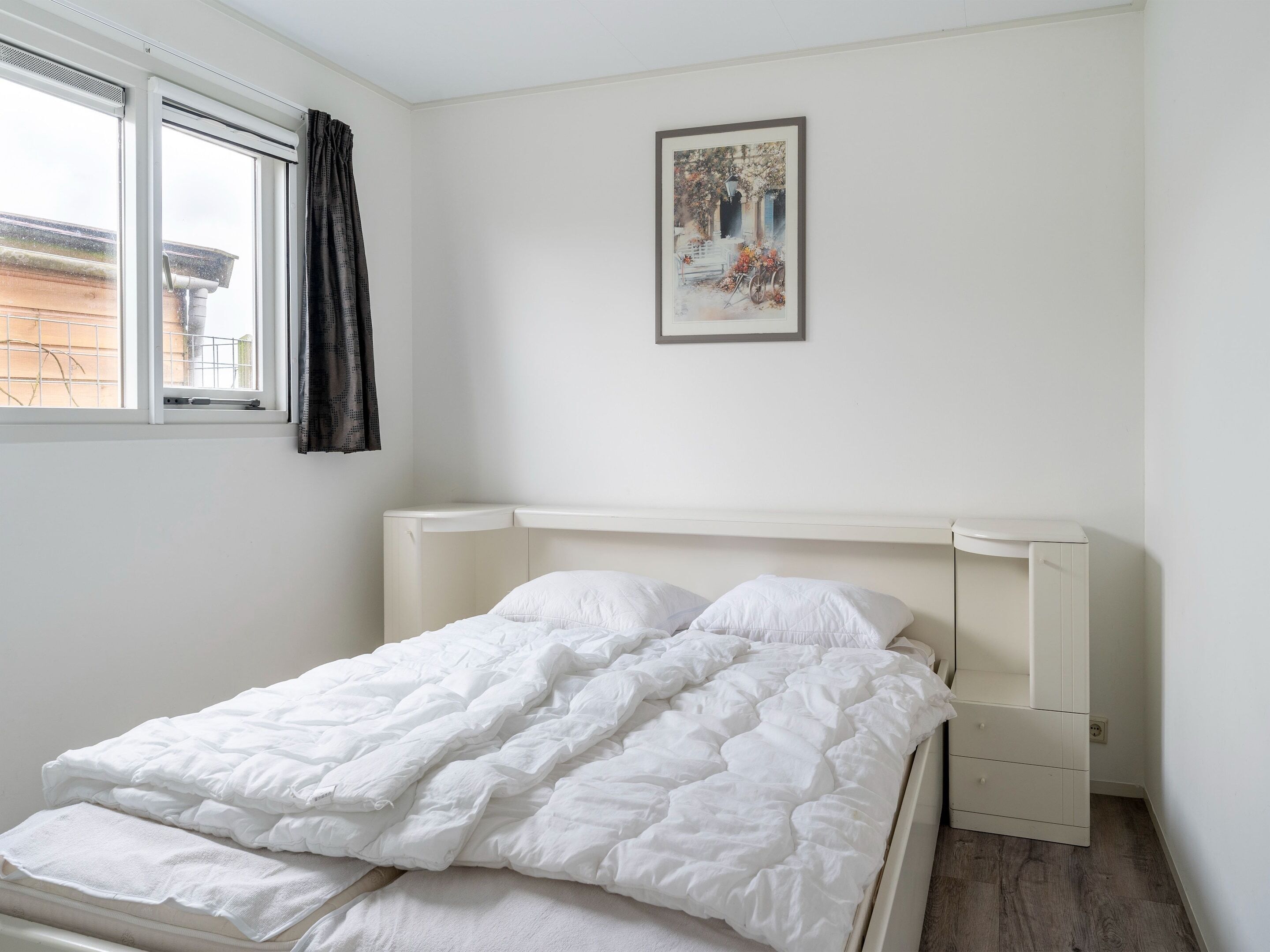Una camera da letto con un letto bianco, una finestra con tende, un quadro appeso al muro e un comodino bianco.