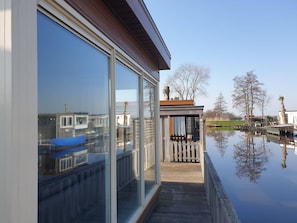 Una casa moderna con amplios ventanales de vidrio, una terraza de madera y vistas a un lago tranquilo con barcos.