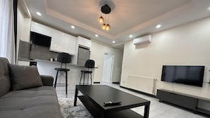 Una sala de estar moderna con un sofá, una mesa de centro y un televisor. Se observa una zona de cocina con armarios y un refrigerador.