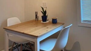 Un coin repas avec une table en bois, deux chaises blanches, une plante en pot et un tableau encadré au mur.