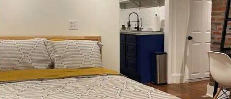 A bedroom with a bed, a small kitchenette, and a door leading to a bathroom.