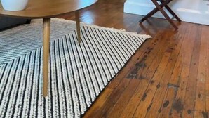 Une pièce avec une table ronde en bois, deux chaises, un tapis à rayures, une grande plante et un tableau abstrait encadré accroché au mur.