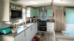 A compact kitchen with a microwave, stove, and sink. There are cabinets, a clock, and a colorful rug. - Chalet in Oostkapelle Near the Beach (Oostkapelle)