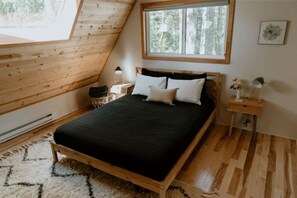 Een slaapkamer met een schuine plafond, een bed met zwart-wit beddengoed, een klein nachtkastje met een lamp en een raam met uitzicht op bomen.
