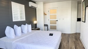 A hotel room with a bed, a nightstand, a lamp, a wardrobe, and a door leading to another room. - Devon Shores - Waterview King Suite (Clearwater)