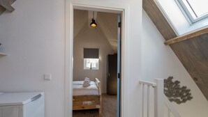 Een smalle gang die leidt naar een slaapkamer met een schuine plafond, een bed, een raam en een plafondlamp.