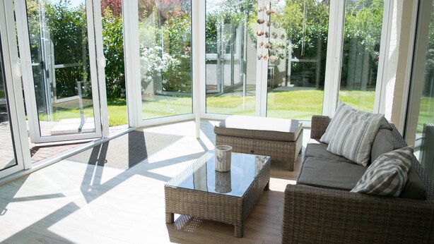 A sunlit room with a glass door leading to a garden, wicker furniture, and a glass coffee table.