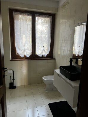 Ein Badezimmer mit Toilette, einem Waschbecken mit schwarzer Schüssel und einem Fenster mit transparenten Vorhängen.