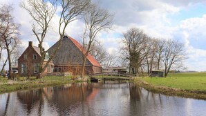A traditional brick house with a red roof, a small bridge over a calm water body, and a grassy field in the background. - Farmhouse in Suwald With Hot Tub (Suwâld)