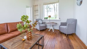 A living room with a brown leather sofa, a glass coffee table, two grey armchairs, a round wooden table, and a window with a view of trees. - Farmhouse in Suwald With Hot Tub (Suwâld)