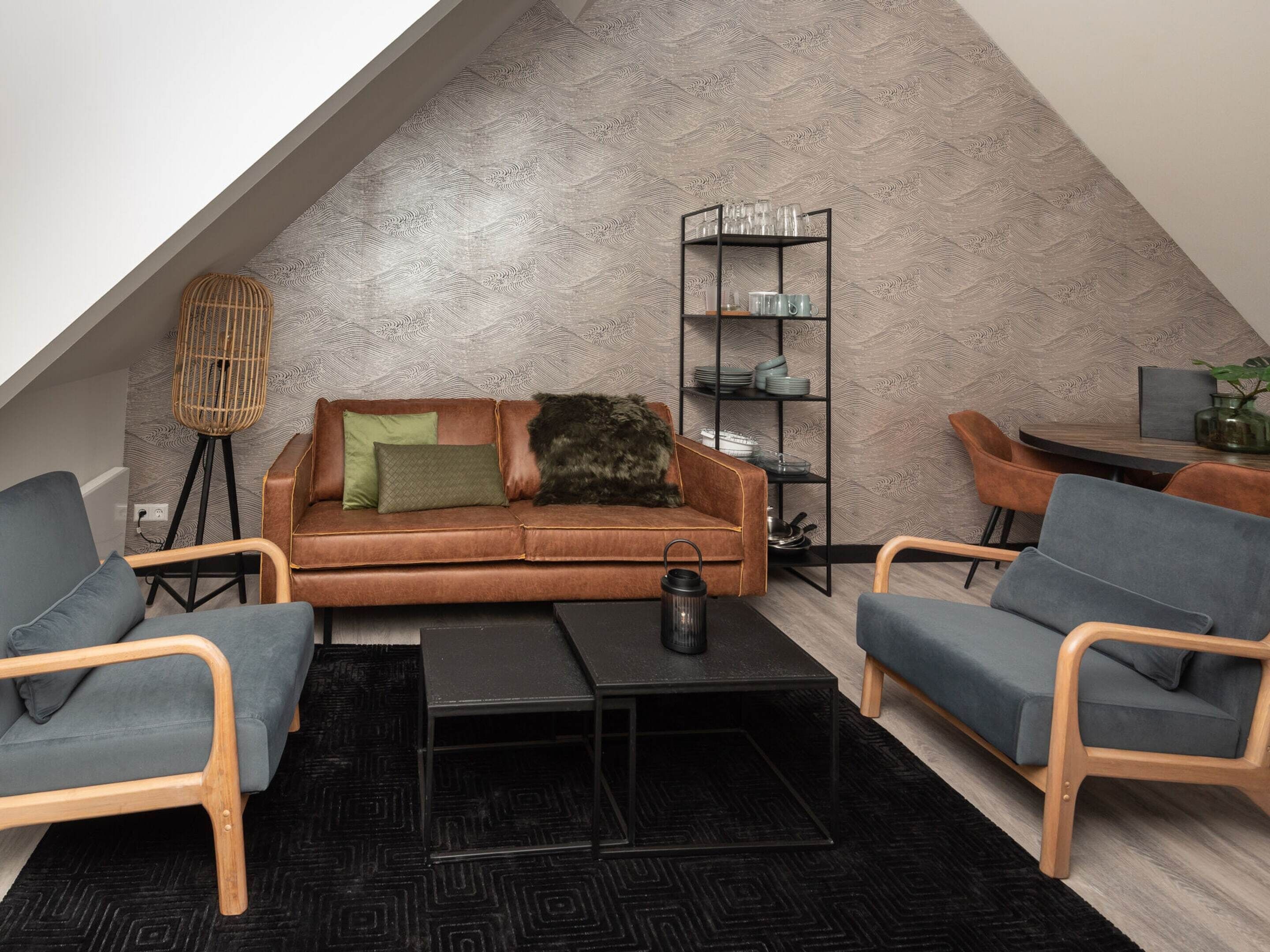 A cozy loft-style living space with a brown leather sofa, two grey armchairs, a black coffee table, and a black rug. A metal shelf unit with dishes and glasses is against the textured wall.