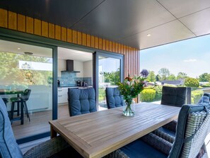 Een moderne buiten eethoek met een houten tafel en stoelen, een glazen wand en uitzicht op de tuin en omliggende huizen.