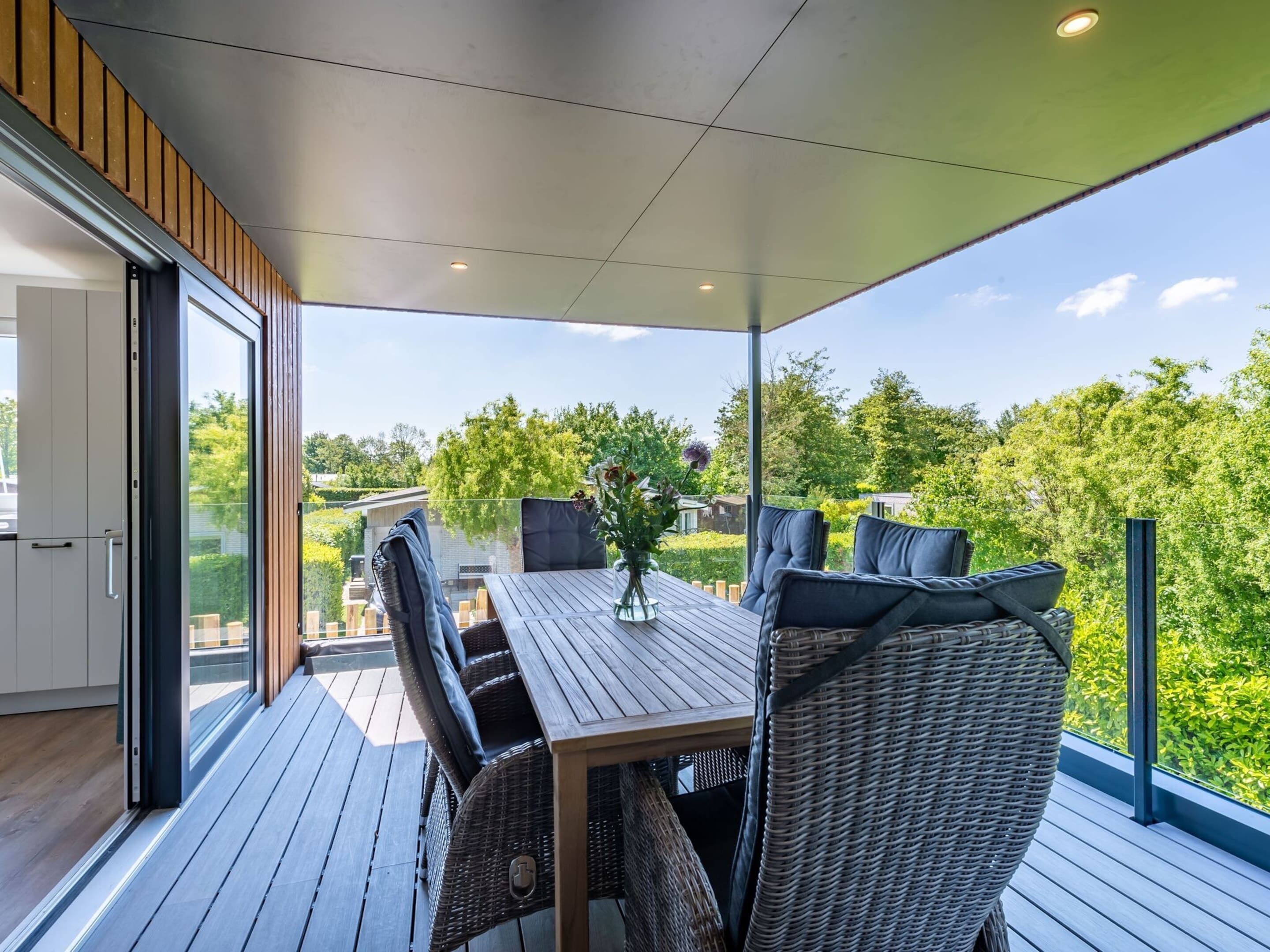 A modern outdoor dining area with a wooden table and wicker chairs, overlooking a lush garden and a clear sky.
