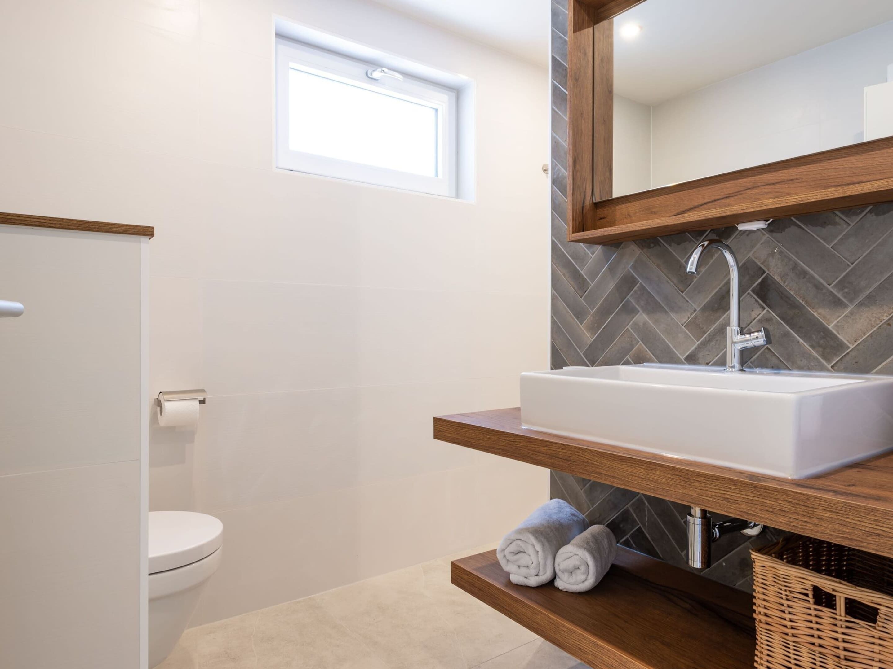 A modern bathroom with a white toilet, a white sink on a wooden shelf, a herringbone-patterned wall, and a small window above the toilet.