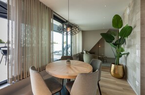 Uma zona de refeições moderna com uma mesa redonda de madeira, cadeiras acolchoadas, um candeeiro pendente e uma grande planta em vaso.