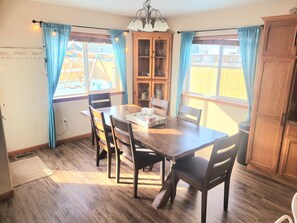 Uma sala de jantar com uma mesa e cadeiras de madeira, um armário com portas de vidro e uma janela com cortinas.