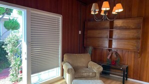 Une pièce dotée d’une porte-fenêtre coulissante, d’un fauteuil beige, d’un pouf assorti et d’un mur en bois avec des étagères intégrées et un lustre.