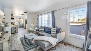 A modern living room with a grey sofa set, a coffee table, and a floor lamp. There is a kitchen area visible in the background. - MB Gem Lower - Steps To The Beach, Ocean Breeze (Manhattan Beach)