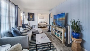 A modern living room with a sofa, coffee table, and a TV. There is a bed in the background and a plant on the side. - MB Gem Lower - Steps To The Beach, Ocean Breeze (Manhattan Beach)