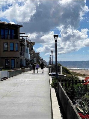 A coastal walkway with modern buildings, a beach view, and street lamps.