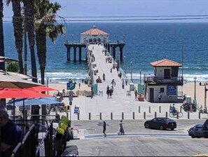A pier extending into the ocean with a building at the end, a beach area with umbrellas, and a small structure on the right.