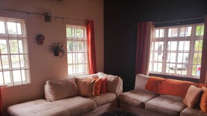 A living room with two beige sofas, a black ottoman, and a window with curtains. - Flagstaff Home Away From Home (Port of Spain)
