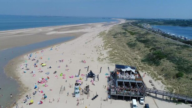 Uma praia com um pavilhão e palco para eventos, rodeada por areia e dunas.