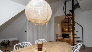 Un coin repas avec une table en bois, des chaises blanches, un luminaire suspendu en fibres tressées et, au fond, un escalier en colimaçon.