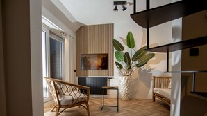 Un salon moderne avec un parquet en chevrons, un fauteuil en osier, une grande plante en pot, un téléviseur à écran plat fixé sur un mur en panneaux de bois, et un escalier avec une rampe noire.