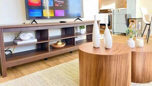 Un salon moderne avec un téléviseur à écran plat fixé sur un meuble TV en bois, une table basse ronde en bois et un coin cuisine à l’arrière-plan.