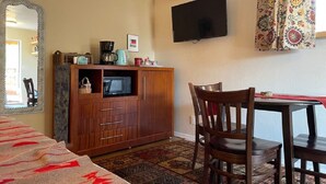 A room with a wooden cabinet, a TV mounted on the wall, a dining table with chairs, and a patterned carpet.