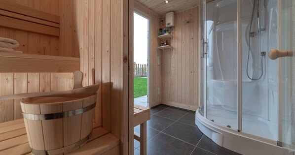 Un sauna en bois avec un seau en bois, un espace douche fermé par des parois de verre et une porte donnant sur l’extérieur.