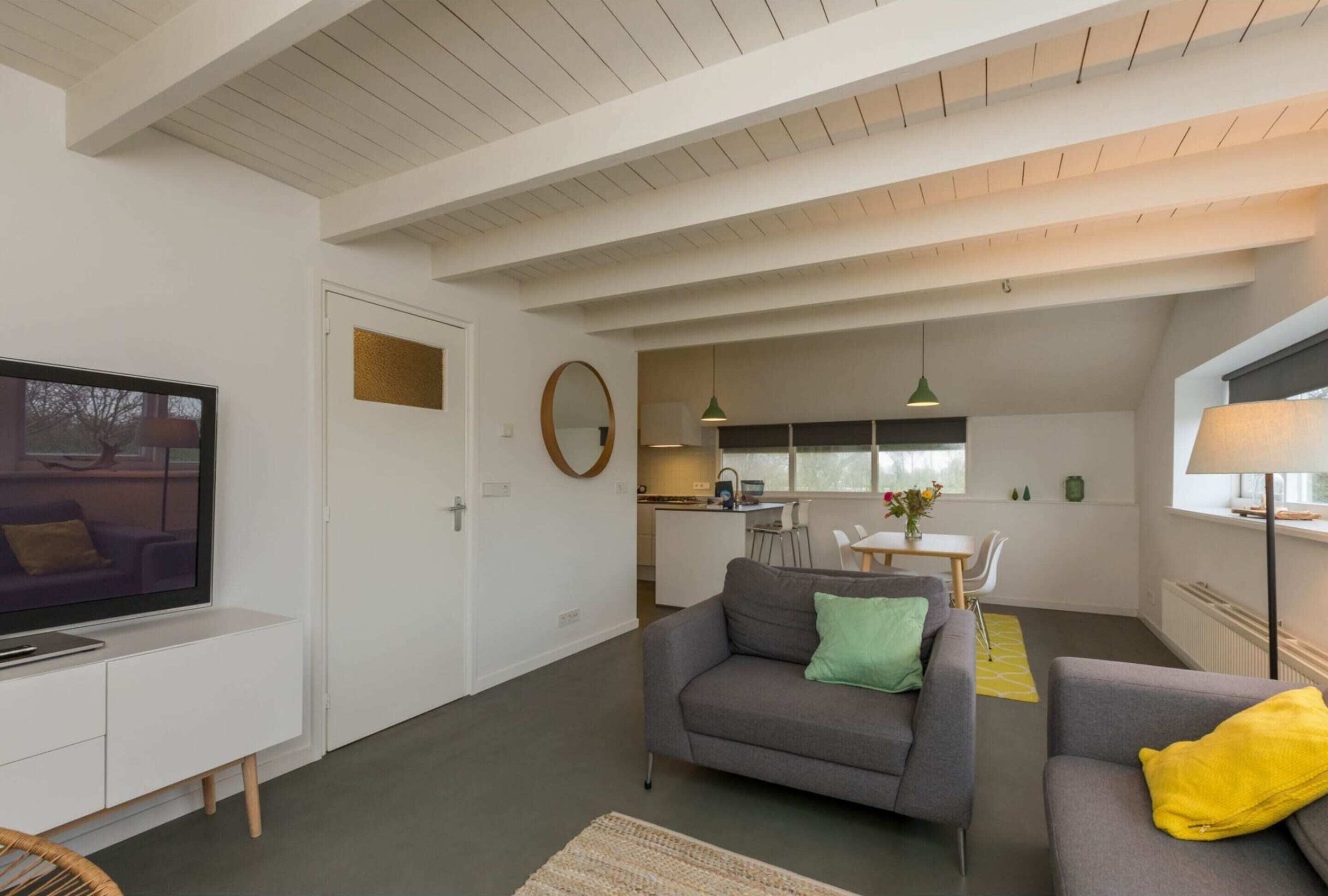 Uma sala de estar moderna com televisão de tela plana, sofás cinzas, um espaço de refeições e uma cozinha ao fundo.