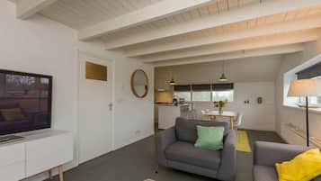 Uma sala de estar moderna com televisão de tela plana, sofás cinzas, um espaço de refeições e uma cozinha ao fundo.