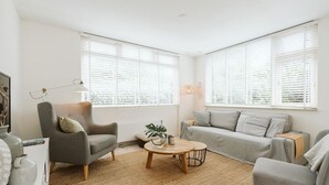 Un salon moderne avec un canapé gris, des fauteuils, une table basse et de grandes fenêtres équipées de stores.